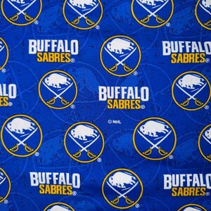 May include: A blue fabric with a repeating pattern of the Buffalo Sabres logo and text. The logo features a white buffalo, crossed swords, and a yellow circle. The text "BUFFALO SABRES" is also present.