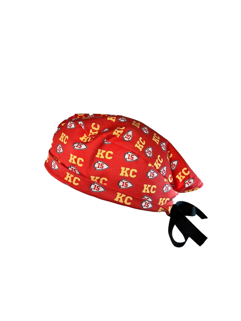 May include: Red and yellow patterned surgical scrub hat with a black tie. The pattern features the letters "KC" and the number "15" repeated throughout.