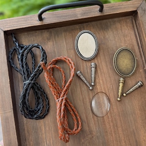 May include: A collection of jewelry making supplies including one black braided cord, one brown braided cord, two oval metal cabochon settings in silver and bronze tones, four matching metal end caps, and one clear oval glass cabochon dome.