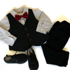 May include: A formal outfit for a child, including a black vest with brown buttons, a striped button-down shirt, a red and black plaid bow tie, black pants, and black sneakers. The ensemble is laid out on a white surface.