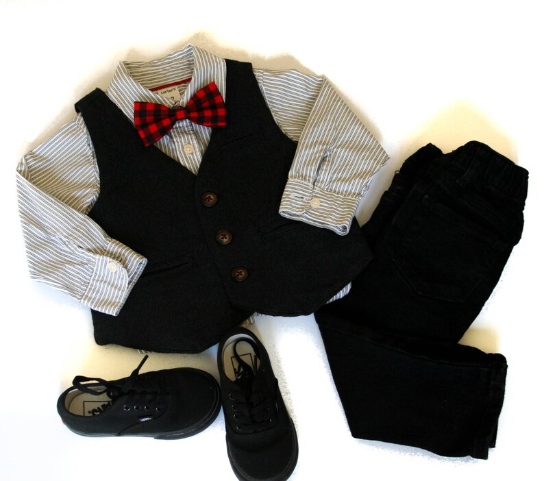 May include: A formal outfit for a child, including a black vest with brown buttons, a striped button-down shirt, a red and black plaid bow tie, black pants, and black sneakers. The ensemble is laid out on a white surface.