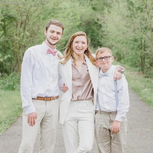 May include: Three people smiling and posing for a photo outdoors. The person on the left is wearing a white button-down shirt, khaki pants, and a pink bow tie. The person in the middle is wearing a white suit and a pink blouse. The person on the right is wearing a white shirt, khaki pants, and a blue bow tie.