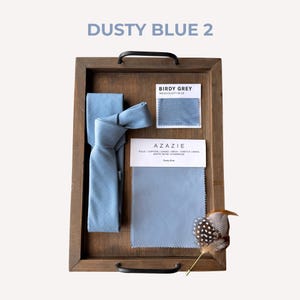 May include: A wooden tray holds a dusty blue tie, fabric swatches, and a decorative feather. The text "DUSTY BLUE 2" is at the top. The fabric swatches are labeled "BIRDY GREY" and "AZAZIE".