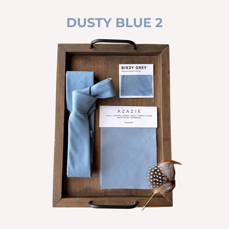 May include: A wooden tray holds a dusty blue tie, fabric swatches, and a decorative feather. The text "DUSTY BLUE 2" is at the top. The fabric swatches are labeled "BIRDY GREY" and "AZAZIE".