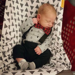 May include: A sleeping baby wearing a gray long-sleeved shirt with a tree pattern, black pants, and a red and black plaid bow tie. The baby is in a shopping cart seat with a gray and white triangle pattern.