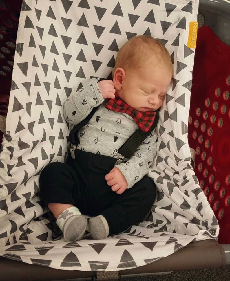 May include: A sleeping baby wearing a gray long-sleeved shirt with a tree pattern, black pants, and a red and black plaid bow tie. The baby is in a shopping cart seat with a gray and white triangle pattern.