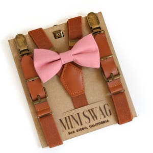 May include: A set of brown suspenders with a pink bow tie, displayed on a cardboard backing. The suspenders have leather accents and metal hardware. The bow tie is made of a solid pink fabric. The text "MINI SWAG SAN DIEGO, CALIFORNIA" is printed on the cardboard.