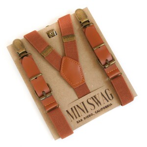 May include: Rust-colored suspenders with brown leather accents and brass-colored hardware. The suspenders are displayed on a cardboard backing with the text "MINI SWAG SAN DIEGO, CALIFORNIA".