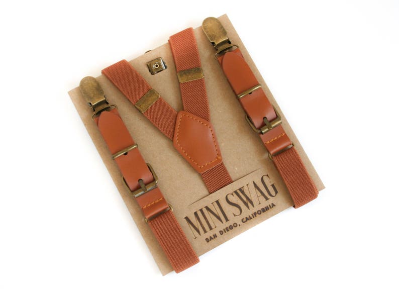May include: Rust-colored suspenders with brown leather accents and brass-colored hardware. The suspenders are displayed on a cardboard backing with the text "MINI SWAG SAN DIEGO, CALIFORNIA".