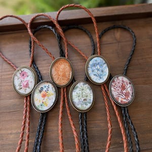 May include: A collection of bolo ties with oval pendants featuring floral designs. The pendants are set in antique brass-colored frames and hang from braided leather cords in brown and black. The floral patterns include pink, orange, blue, and green.