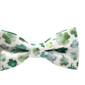May include: A bow tie with a white background and a pattern of green shamrocks. The shamrocks vary in shades of green, creating a watercolor effect. The bow tie is tied in the center and is ready to wear.