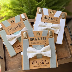 May include: Three sets of suspenders and bow ties on cardboard backing. Each set includes a bow tie with a cross pattern, suspenders, and a name tag. The names "Ethan", "Adam", and "David" are printed on the tags, along with the text "Mini Swag".