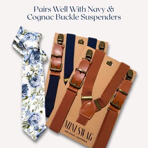 May include: A floral tie with blue roses and a pair of navy and cognac buckle suspenders. The suspenders have leather accents and metal hardware. The text "Pairs Well With Navy & Cognac Buckle Suspenders" is at the top.