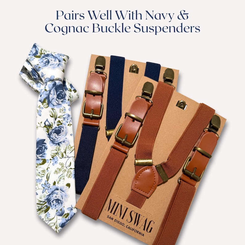 May include: A floral tie with blue roses and a pair of navy and cognac buckle suspenders. The suspenders have leather accents and metal hardware. The text "Pairs Well With Navy & Cognac Buckle Suspenders" is at the top.