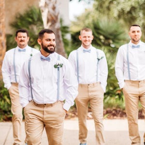 May include: Four people wearing white button-down shirts, khaki pants, light blue bow ties, and light blue suspenders. Each person has a small flower pinned to their shirt. The group is standing outside in front of green foliage.