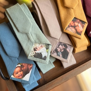 May include: An assortment of neckties in shades of blue, green, beige, and mustard. Each tie has a personalised photograph printed on the front. The ties are displayed on a wooden tray, highlighting the variety of designs and colours.
