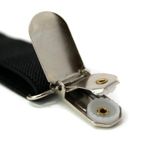 May include: Close-up of a black suspender strap with a silver-colored metal clip. The clip has a white plastic insert with a gold-colored center. The suspender strap is made of a woven material.