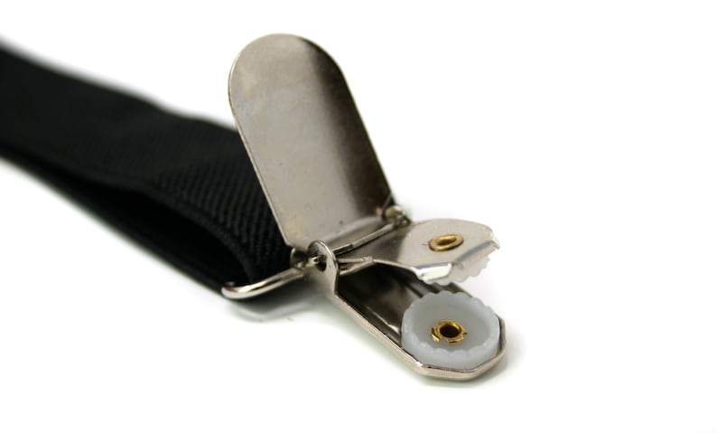 May include: Close-up of a black suspender strap with a silver-colored metal clip. The clip has a white plastic insert with a gold-colored center. The suspender strap is made of a woven material.