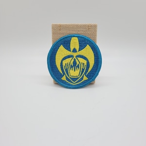 May include: Round embroidered patch with a blue border and a yellow emblem. The emblem features a stylized design with a central figure and wing-like shapes. The patch is displayed on a small burlap-like square against a white background.