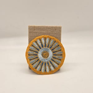 May include: A round, embroidered patch with a floral design. The patch features a yellow border with blue and light blue petal-like shapes radiating from a light blue center. The patch is displayed on a small, tan, rectangular block.