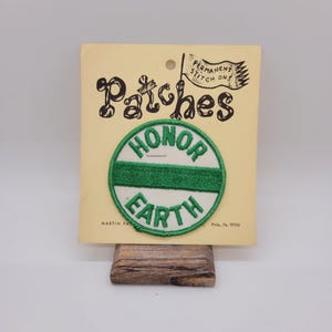 May include: A green and white round iron-on patch with the words "HONOR EARTH" in a circle. The patch is on a yellow card with the words "Patches" and "Permanent Stitch Ons" printed on it.