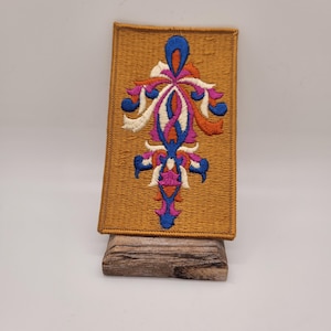 May include: A gold embroidered patch with a colorful, intricate design. The design features a central motif with swirling lines and shapes in shades of blue, pink, orange, and white.