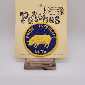 May include: A round patch with a yellow pig illustration on a blue background, encircled by a yellow border. The words "PRIDE", "INTEGRITY", and "GUTS" are displayed. The patch is attached to a cream-colored card with the word "Patches" and "PERMANENT STITCH ONS" printed on it.
