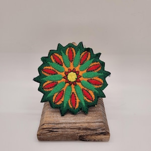 May include: A green, orange, and red embroidered flower patch with a yellow center. The patch is on a wooden stand.