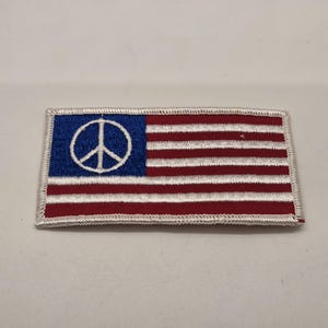 May include: Embroidered patch featuring the American flag with a peace symbol replacing the stars. The patch has a blue field with a white peace symbol and red and white stripes. The patch is rectangular with a white border.