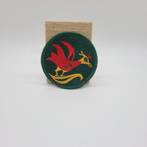 May include: A green circular patch with an embroidered red and yellow bird design. The bird has a long neck and a long tail.