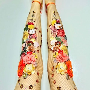 Könnte beinhalten: Beine in hautfarbenen Fischstrumpfhosen, verziert mit bunten Blumenverzierungen und glitzernden Edelsteinen. Die Füße stecken in orangefarbenen Plateau-Heels mit geometrischem Muster. Die Beine sind nach oben gerichtet.