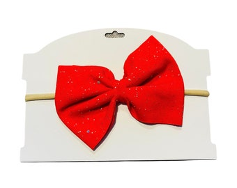 Può includere: Fascia per capelli con fiocco rosso glitterato su una carta bianca. Il fiocco è fatto di tessuto rosso con glitter argentati.