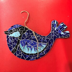 Puede incluir: Adorno de pájaro de mosaico en tonos azules, negros y celeste, con una cuerda de yute para colgar. El pájaro está hecho de pequeñas baldosas de forma irregular, creando una superficie texturizada. El adorno está sobre un fondo rojo.