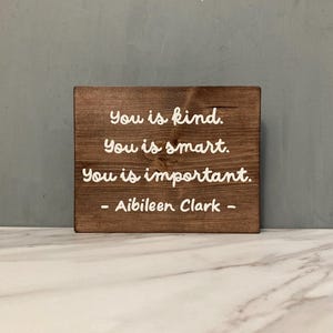 May include: A wooden sign with the words "You is kind. You is smart. You is important." in white script. The sign is signed "- Aibileen Clark -" and is a decorative item.