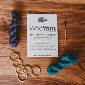 May include: A Dorset button making kit with two skeins of yarn, one dark blue and one teal green, six gold rings, a needle, and instructions on a white card with a sheep logo and the text "WeeYarn Designs".