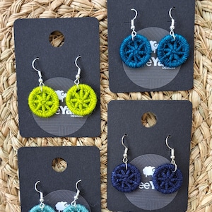 Könnte beinhalten: Vier Paar handgefertigte Ohrringe mit Knopfdesign. Die Ohrringe werden auf schwarzen Karten mit dem Text "Crafty Yarn Designs" präsentiert. Die Ohrringe sind in den Farben Türkis, Grün und Blau gehalten.
