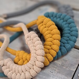Peut inclure: Trois colliers en corde tressée de couleur beige, jaune moutarde et bleu sarcelle. Les colliers sont disposés en pile, le collier beige en bas et le collier sarcelle en haut.