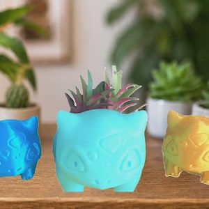 May include: Three Bulbasaur-shaped planters in blue, teal, and gold. The teal planter holds small succulents. The planters are on a wooden surface with potted plants in the background. A fun and unique home decor item.