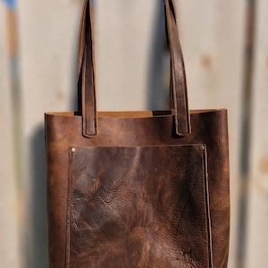 May include: A brown leather tote bag with a front pocket and two handles. The bag has a textured surface and a small dog silhouette on the front pocket. The handles are attached with brass rivets. The bag is designed for carrying items.
