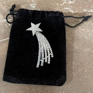 May include: A silver-toned starburst brooch with a five-pointed star and a trailing design of sparkling crystals. The brooch is displayed on a black velvet drawstring bag.