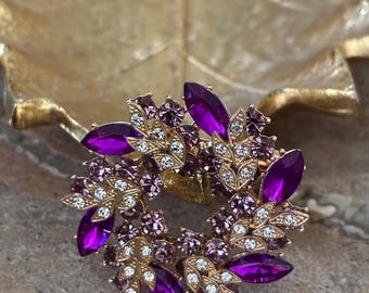 May include: A gold-toned brooch with a wreath design. The wreath is adorned with large, deep purple, oval-shaped gemstones and smaller purple and clear crystals. The brooch has a detailed, floral-inspired pattern.