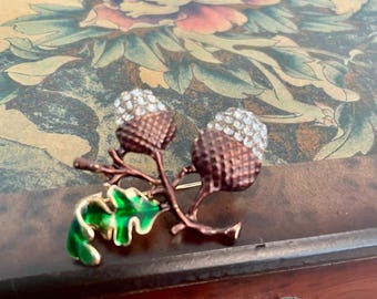 May include: A gold-toned acorn brooch with two acorns and a green leaf. The acorns are decorated with clear rhinestones.