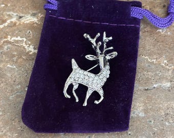 May include: A silver-toned deer brooch featuring a polished metal finish on the head and antlers, with the body encrusted in clear, sparkling rhinestones. The brooch is positioned on a small, rectangular purple velvet drawstring pouch.