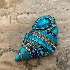Könnte beinhalten: Türkisfarbene Muschelbrosche mit einem großen tropfenförmigen blauen Edelstein oben. Die Brosche ist mit blauen und klaren Kristallen in verschiedenen Größen verziert, die einen funkelnden Effekt erzeugen. Das Design hat ein geschichtetes, strukturiertes Aussehen.