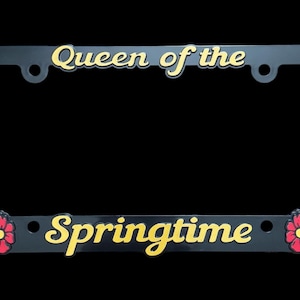 May include: Black license plate frame with gold lettering that reads "Queen of the Springtime". The frame is decorated with red flowers.