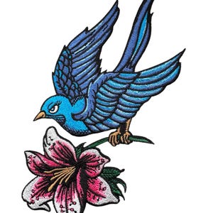 May include: An embroidered patch depicting a blue bird perched on a pink lily flower. The bird is intricately designed with blue and black feathers. The lily flower has pink petals with white accents.