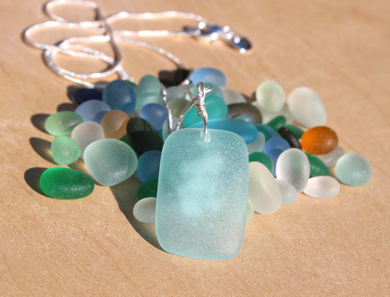 May include: A silver chain necklace with a light blue sea glass pendant. The pendant is surrounded by a variety of sea glass pieces in shades of blue, green, and white.
