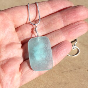 May include: A silver chain necklace with a light blue sea glass pendant. The pendant is rectangular and has a frosted finish.