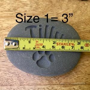 May include: A gray stone with the name "Tillie" engraved on it, along with a paw print. A measuring tape shows the stone is 3 inches in size.  Size 1 = 3"