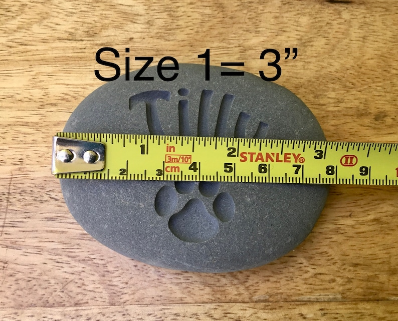 May include: A gray stone with the name "Tillie" engraved on it, along with a paw print. A measuring tape shows the stone is 3 inches in size.  Size 1 = 3"